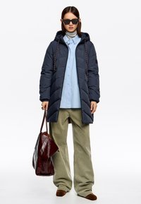 Kobieta w okularach przeciwsłonecznych, długim, ciemnym pikowanym płaszczu, jasnoniebieskiej koszuli, luźnych zielonych spodniach, brązowych butach, trzymająca dużą ciemnoczerwoną torbę.