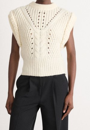 Gilet sans manches en maille crème avec motifs torsadés et côtes, porté avec un pantalon noir taille haute sur une personne à la peau foncée.