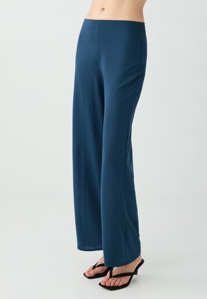Person trägt hoch taillierte, weit geschnittene marineblaue Hose und schwarze offene Sandalen mit niedrigen Absätzen, steht vor hellem Hintergrund.