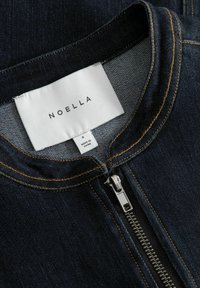 Ciemna kurtka dżinsowa z zamkiem błyskawicznym, zaokrąglonym kołnierzem i kontrastującym pomarańczowym przeszyciem. Metka pokazuje "NOELLA", rozmiar S i "Made in China".