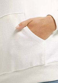 Biała bluza z długim rękawem z boczną kieszenią, z widoczną ręką spoczywającą w środku. Miękka tekstura, ściągacz na dole, minimalistyczny design.