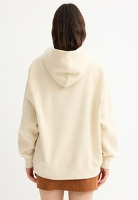 Sudadera beige de gran tamaño con capucha ajustable, mangas raglán y puños acanalados, combinada con una falda marrón con textura.