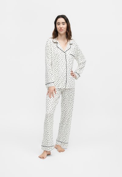 Γυναίκα που στέκεται ξυπόλητη, φοράει λευκή πιτζάμα με μακριά μανίκια και κουμπιά, με μαύρα σχέδια καρδιών και μαύρα διακοσμητικά ρέλια.