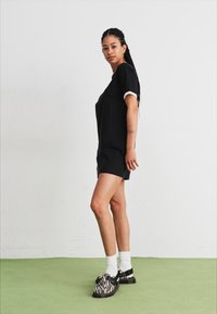 Μαύρο φόρεμα με κοντά μανίκια και λευκές μανσέτες, συνδυασμένο με παπούτσια με σχέδιο ζέβρας και λευκές κάλτσες, σε ανοιχτό φόντο με πράσινο δάπεδο.