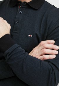 Sötétkék pólóing, textúrált anyagból készült, fekete gallérral, gombos kivágással és kis logóval, piros, fehér és kék színekkel.