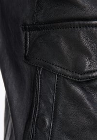 Nahaufnahme einer schwarzen Lederjacke, die detaillierte Nähte, eine strukturierte Oberfläche und eine aufgesetzte Tasche mit sichtbarem Beschlag zeigt.
