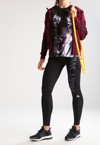 Burgundy vetoketjullinen huppari, musta graafinen tiikeri-printti t-paita, mustat leggings valkoisella tekstillä ja mustat lenkkarit. Pitää kädessään keltaista jumppakehää.