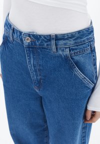 Jeans de mezclilla azul con un corte relajado. Las características incluyen un cierre de botón, cinco bolsillos y costuras en naranja contrastante a lo largo de las costuras.