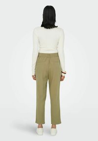 Une personne se tient de dos, portant un haut à manches longues blanc et un pantalon vert olive, avec des chaussures blanches, devant un fond clair et uni.