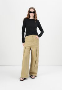 Zwarte long sleeve top, tan brede cargobroek met meerdere zakken, en zwarte flip-flops, tegen een effen witte achtergrond.