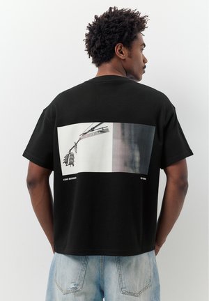 Mężczyzna z kręconymi włosami, ubrany w czarną koszulkę z tyłem ozdobionym zdjęciem sygnalizacji świetlnej i tabliczką ulicy E 23 St w Soho, Nowy Jork.