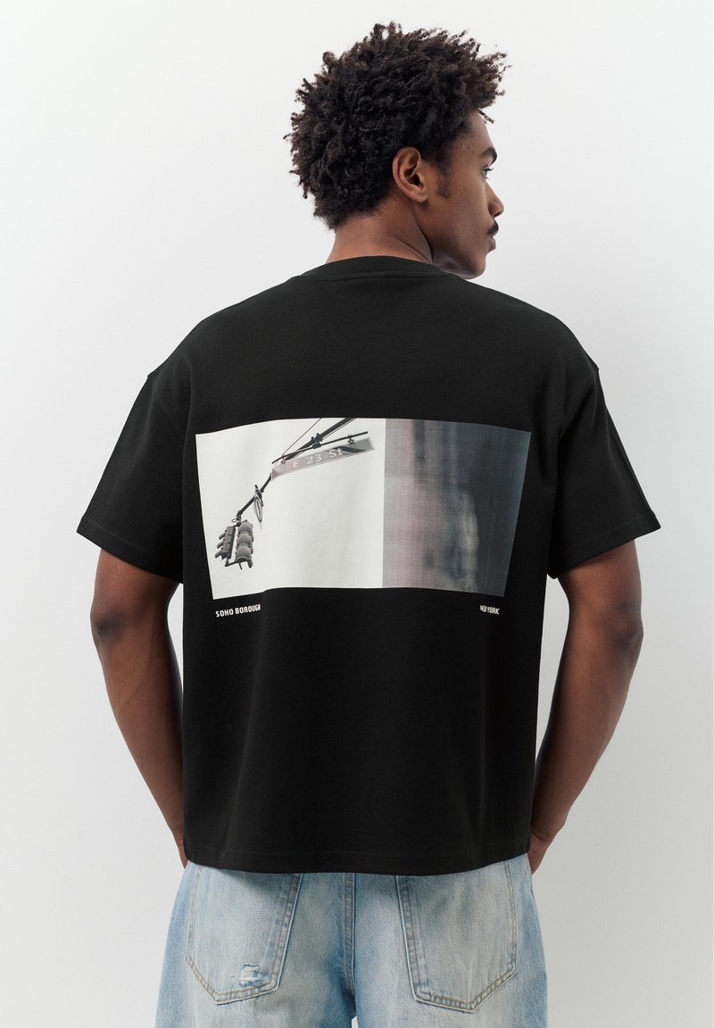 Mann mit lockigem Haar trägt ein schwarzes T-Shirt mit einem Bild einer Ampel und einem Straßenschild für die E 23 St in Soho, New York, auf dem Rücken.