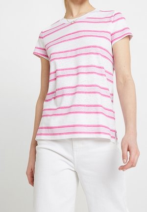Femme portant un t-shirt blanc à manches courtes avec des rayures horizontales roses et un pantalon blanc sur un fond uni.