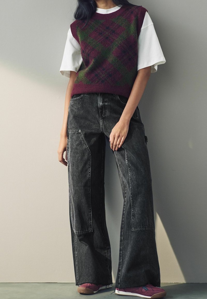 Personne portant une chemise blanche à manches courtes sous un gilet à losanges violet et vert, un jean noir à jambes larges et des baskets violettes, debout contre un mur.