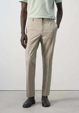 Homme portant un pantalon beige, une chemise de couleur claire et des chaussures en cuir noir, debout devant un fond uni.
