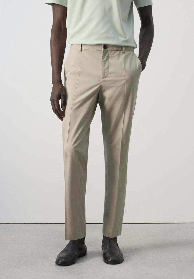 Homme portant un pantalon beige, une chemise de couleur claire et des chaussures en cuir noir, debout devant un fond uni.
