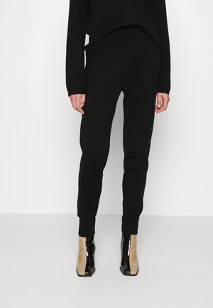 Personne portant un pantalon noir ajusté et un haut à manches longues noir, assortis à des bottines bicolores noires et beiges.