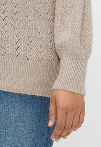 Lys beige strikket genser med et chevronmønster, ribbede mansjetter og en myk tekstur. Hånd synlig, iført denim jeans.
