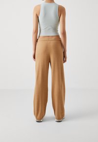 Person wearing a light gray sleeveless top and loose, camel-colored pants, standing facing away on a plain white background.