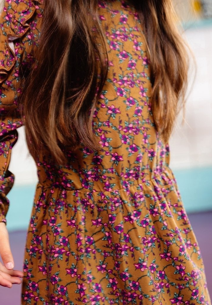 Jeune fille aux longs cheveux bruns portant une robe moutarde à motifs floraux roses avec des manches à volants, debout à l'intérieur.
