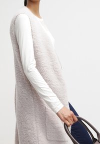 Ljust rosa, texturerad faux fur-väst med öppen front och sidofickor, bärs över en långärmad vit topp, hålls med ett mörkt handtag.