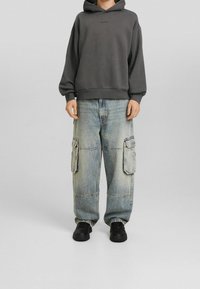 Felpa grigia con cappuccio e un logo discreto, abbinata a jeans cargo blu chiaro con tasche laterali e una vestibilità morbida. Sneakers nere.