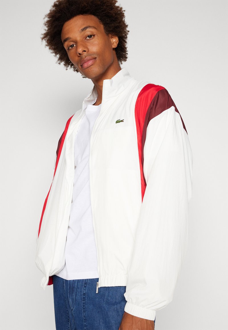 Veste légère blanche avec des accents rouges et bordeaux, fermeture éclair à l'avant, poches latérales et poignets élastiques, présentant un petit logo sur la poitrine.