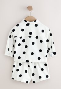 Ensemble chemise à manches longues blanche et short avec de gros pois noirs, suspendu sur un cintre en bois sur un fond clair.