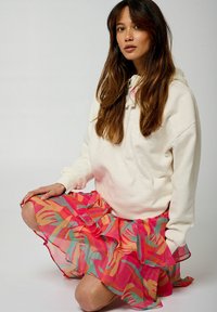 Hoodie crème avec logo texturé, associé à une mini-jupe volantée lumineuse et imprimée en rose, orange et turquoise, fabriquée dans un tissu léger.