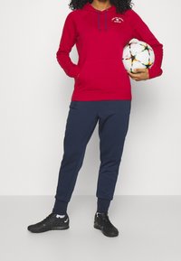 Červená mikina s kapucí, přední kapsou a šňůrkami, v kombinaci s tmavě modrými tepláky. Černé sportovní boty. Drží vícebarevný fotbalový míč.