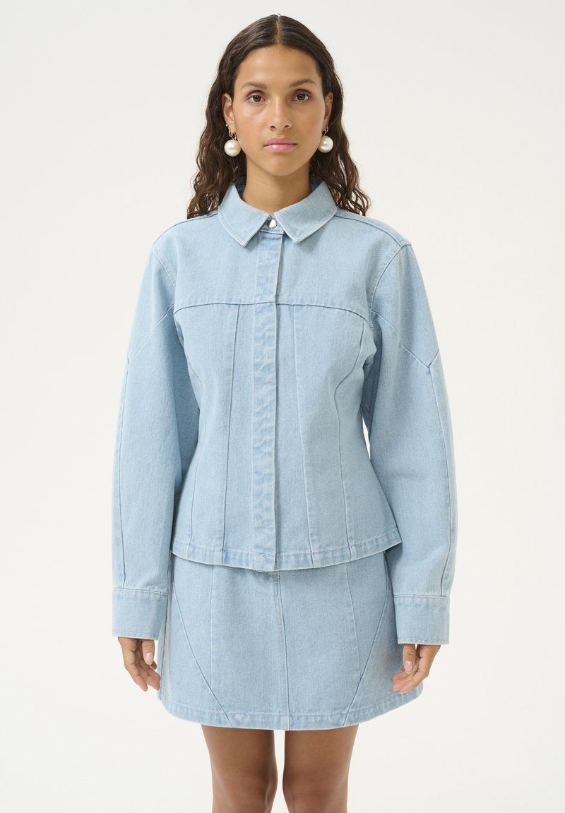 Giacca in denim azzurro chiaro con colletto a punta, design strutturato e maniche lunghe; abbinata a una gonna in denim coordinata con dettagli delle cuciture.