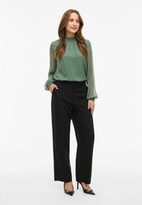Grøn bluse med høj hals i gennemsigtigt stof og flæset manchetter; sorte, skræddersyede bukser med lige pasform; sorte hæle med gyldne detaljer.