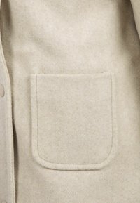 Bežinis vilnonis paltas su apvaliais kampais ir matoma siūle, kairėje pusėje esanti priekinė mygtukų uždarymo dalis.