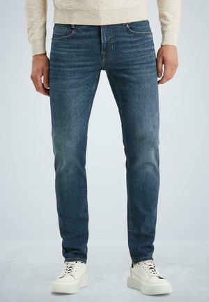 Blue denim jeans with a slim fit, subtle whiskering, and five-pocket design. Paired with white sneakers, featuring minimal detailing.