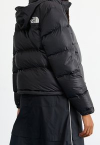 Svart puffjacka med huva, med kviltad vaddering, elastiska ärmar och The North Face-logotyp på axeln. Buren över ett svart plagg.