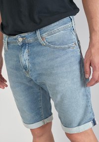 Shorts en denim bleu clair avec ourlets roulés, design à cinq poches et décoloration subtile. Fermeture à bouton en métal et détails de couture visibles.
