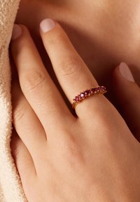 Goldring, verziert mit fünf runden, tiefrosa Steinen, in einem glatten, glänzenden Band gefasst, getragen an einer Hand mit heller Haut und neutralem Nagellack.