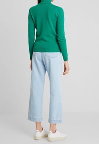 Maglione green di collo alto realizzato in morbido tessuto a maglia, abbinato a jeans a gamba larga blu chiaro e scarpe da ginnastica bianche con discreto marchio sul tallone.