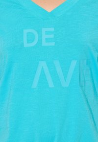 Camisa de cuello en V turquesa con el texto "MONTAR LA OLA" en diferentes tamaños de fuente, con una textura suave y una tela ligera.