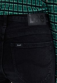 Jeans de mezclilla negra con una cinturilla texturizada y una etiqueta de cuero con el logo de "Lee". El bolsillo lateral incluye una pequeña etiqueta de tela.