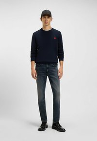 Maglione navy con logo rosso, indossato con jeans blu sbiaditi e scarpe nere stringate, su uno sfondo grigio semplice.