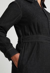 Černé riflové šaty s límečkem košile, dlouhými rukávy a pasem se zavazováním. Jsou zdobeny kontrastním žlutým šitím a mají boční kapsy.