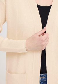 Gilet en tricot beige clair avec un col en V, une ouverture à l'avant et des manches longues. Comprend des poches et un tissu doux et texturé.