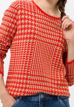 Femme portant un pull à motif pied-de-poule rouge et beige avec col rond et un jean bleu, tenant une tresse avec la main droite.