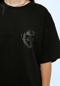 Zwarte t-shirt met korte mouwen van katoen, met een opvallend zwart vlamlogo op de linkerborst. Gladde textuur.