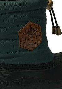 Botte imperméable avec une tige en tissu vert foncé, une semelle en caoutchouc noir texturée et un patch en cuir marron avec un dessin de montagne et un logo.
