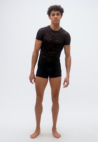 Νεαρός άνδρας στέκεται ξυπόλυτος φορώντας μαύρο διαφανές δαντελωτό μπλουζάκι και μαύρα στενά σορτς σε απλό λευκό φόντο.