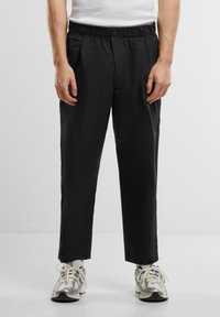 Pantaloni negri cu o croială confortabilă, talie elastică și două buzunare frontale, fabricați dintr-un material neted, purtați cu pantofi sport.