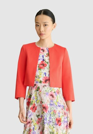 Mujer que lleva una chaqueta corta roja sobre un vestido sin mangas con flores rosas, moradas y amarillas sobre un fondo claro.