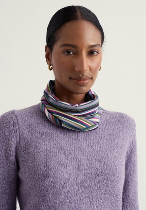 Donna con orecchini a cerchio dorati che indossa un maglione viola lavorato a maglia e una sciarpa a righe multicolori al collo, in posa davanti a uno sfondo neutro.